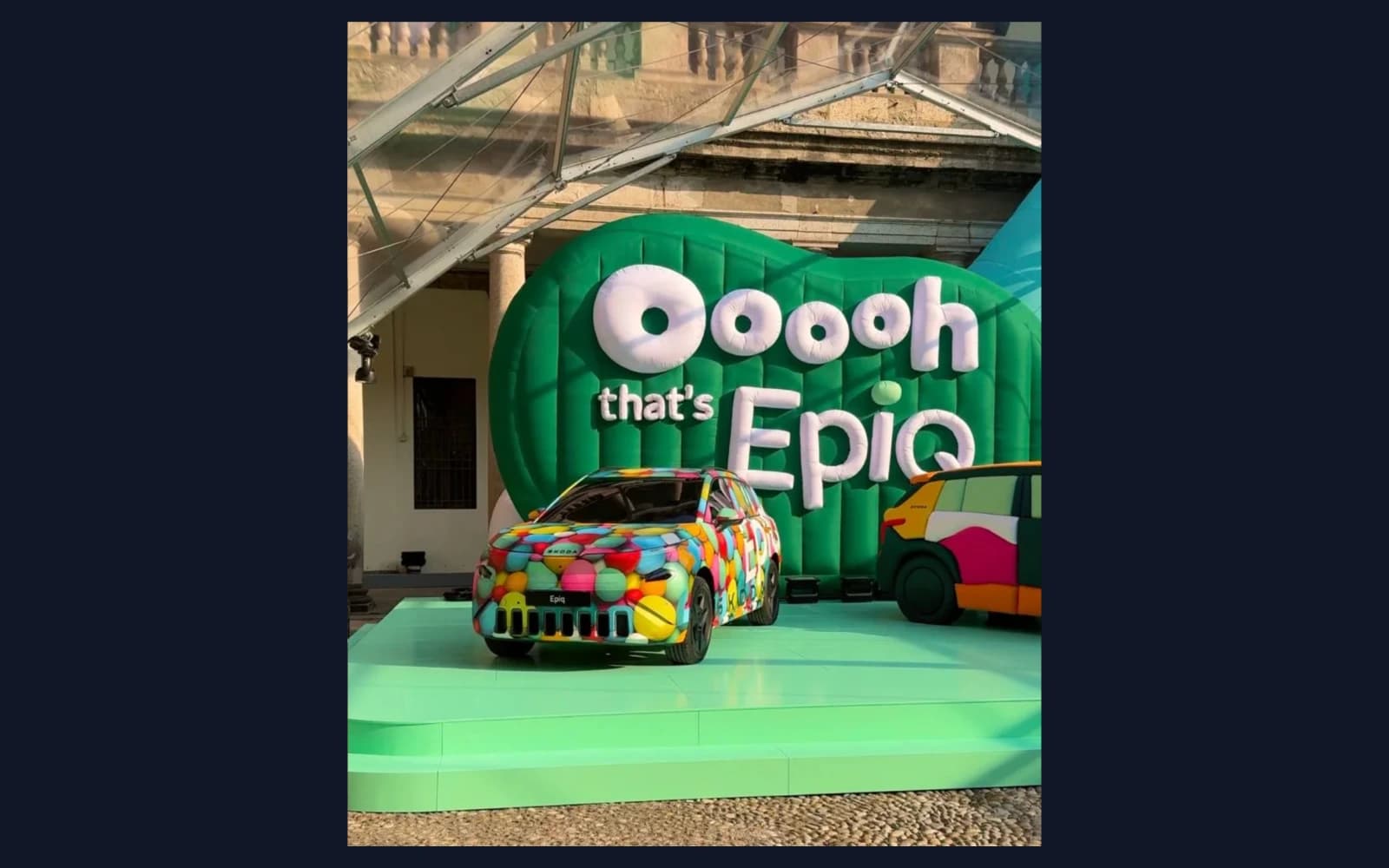 The Škoda EPIQ, wrapped in a colourful pattern, on a mint-green platform in front of a large green inflatable backdrop reading 'Ooooh that's EpiQ' at Milan Design Week.
