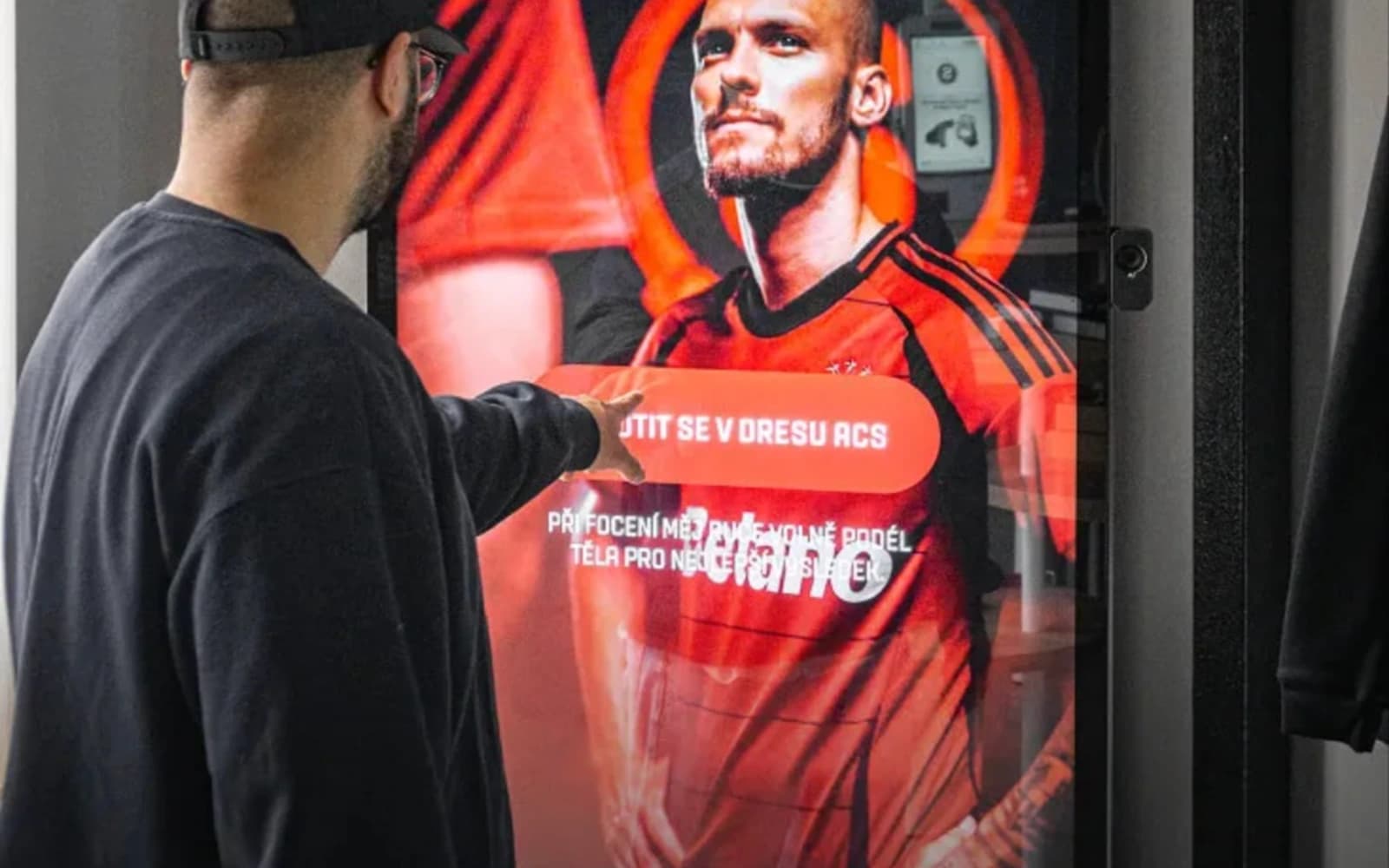 A customer in a black cap reaching out to a large touchscreen in the Sparta Prague Boutique, showing a player in the club's red and black kit with ČSOB and Mastercard logos.